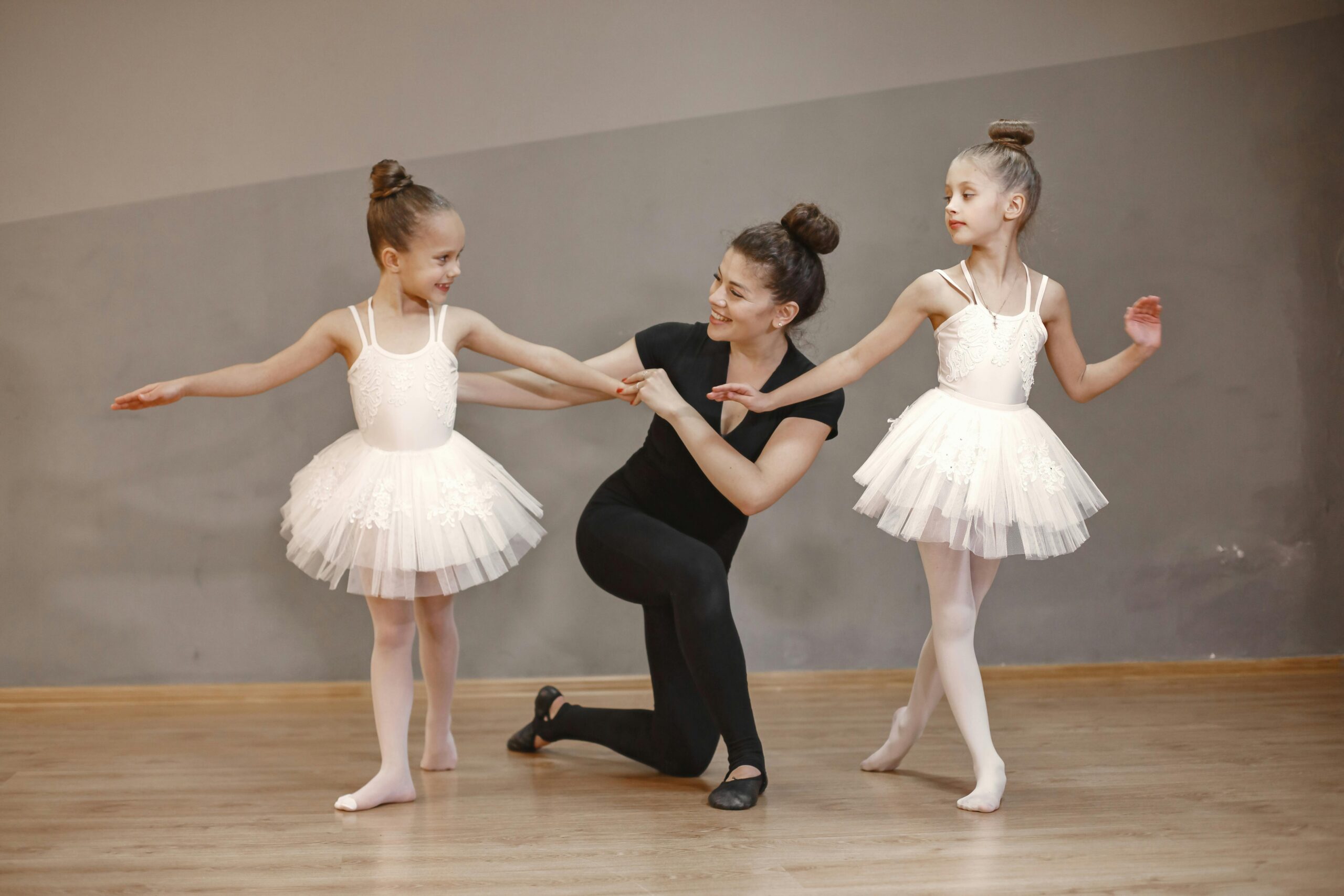 Young girls in tutus being guided by their ballet teacher in a dance studio.