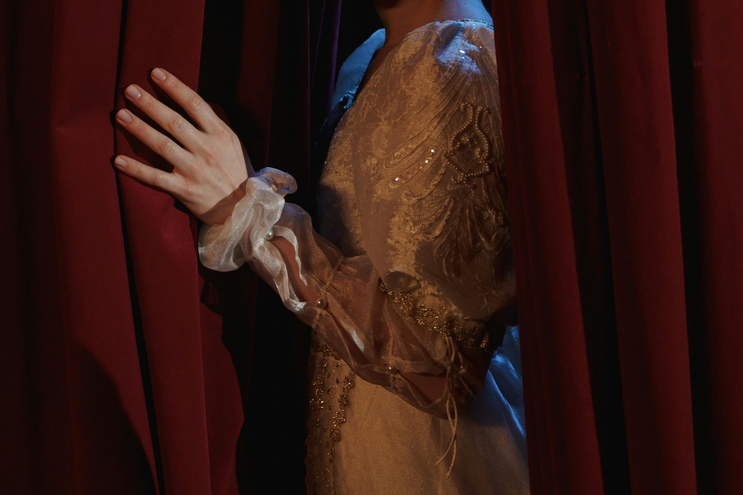 An actress in a vintage costume gracefully pulls back a red curtain on stage, creating a dramatic scene.
