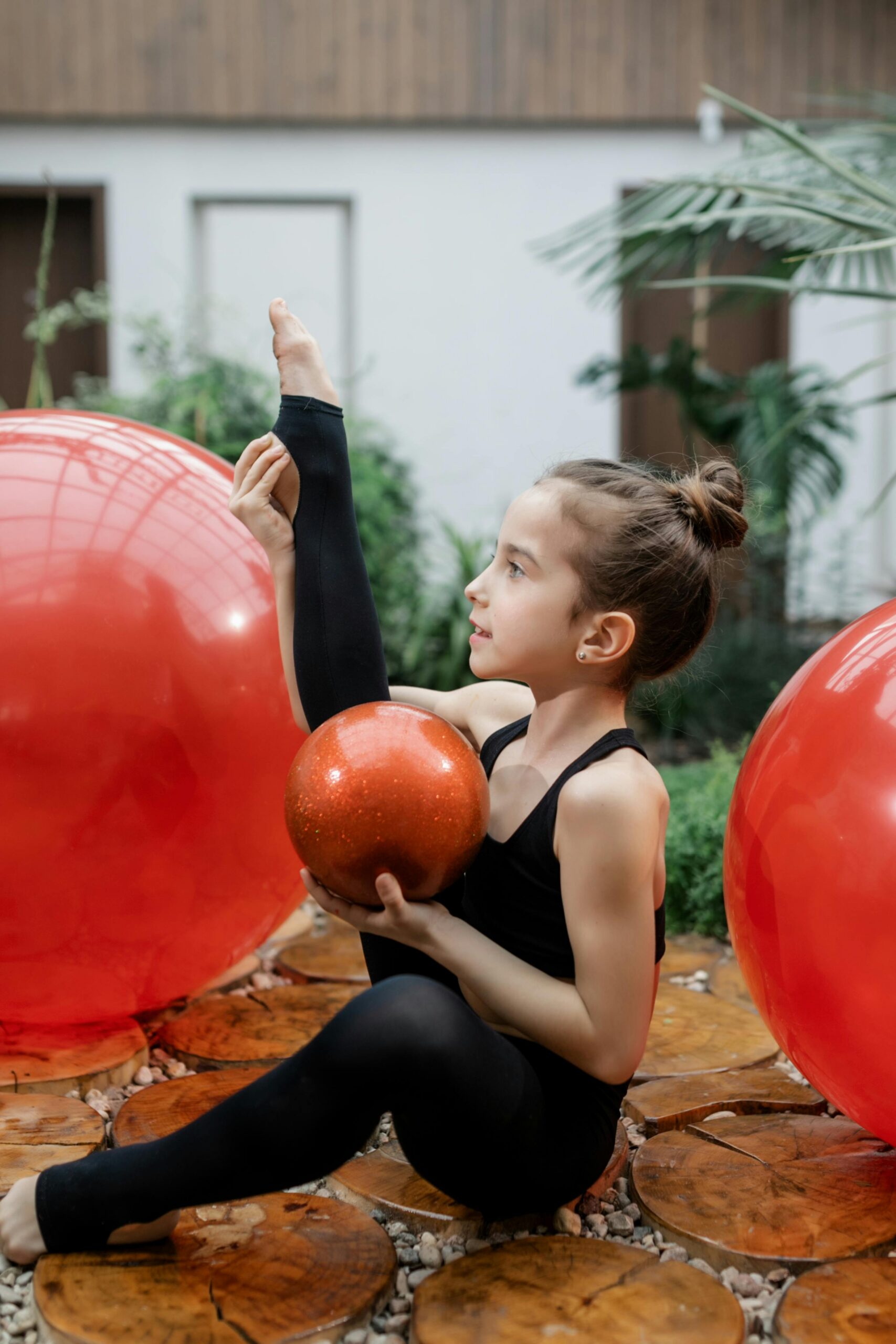 A young girl engages in rhythmic gymnastics indoors, showcasing flexibility with a red ball.