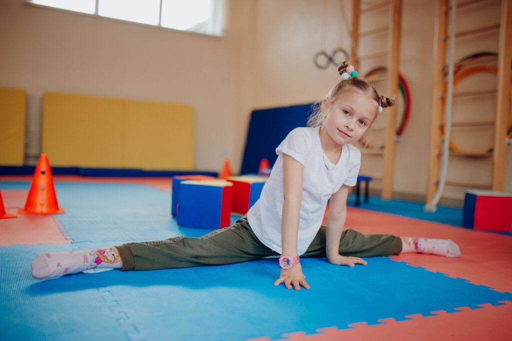 Young girl showing flexibility while practicing splits indoors on colorful mats in a playroom setting.