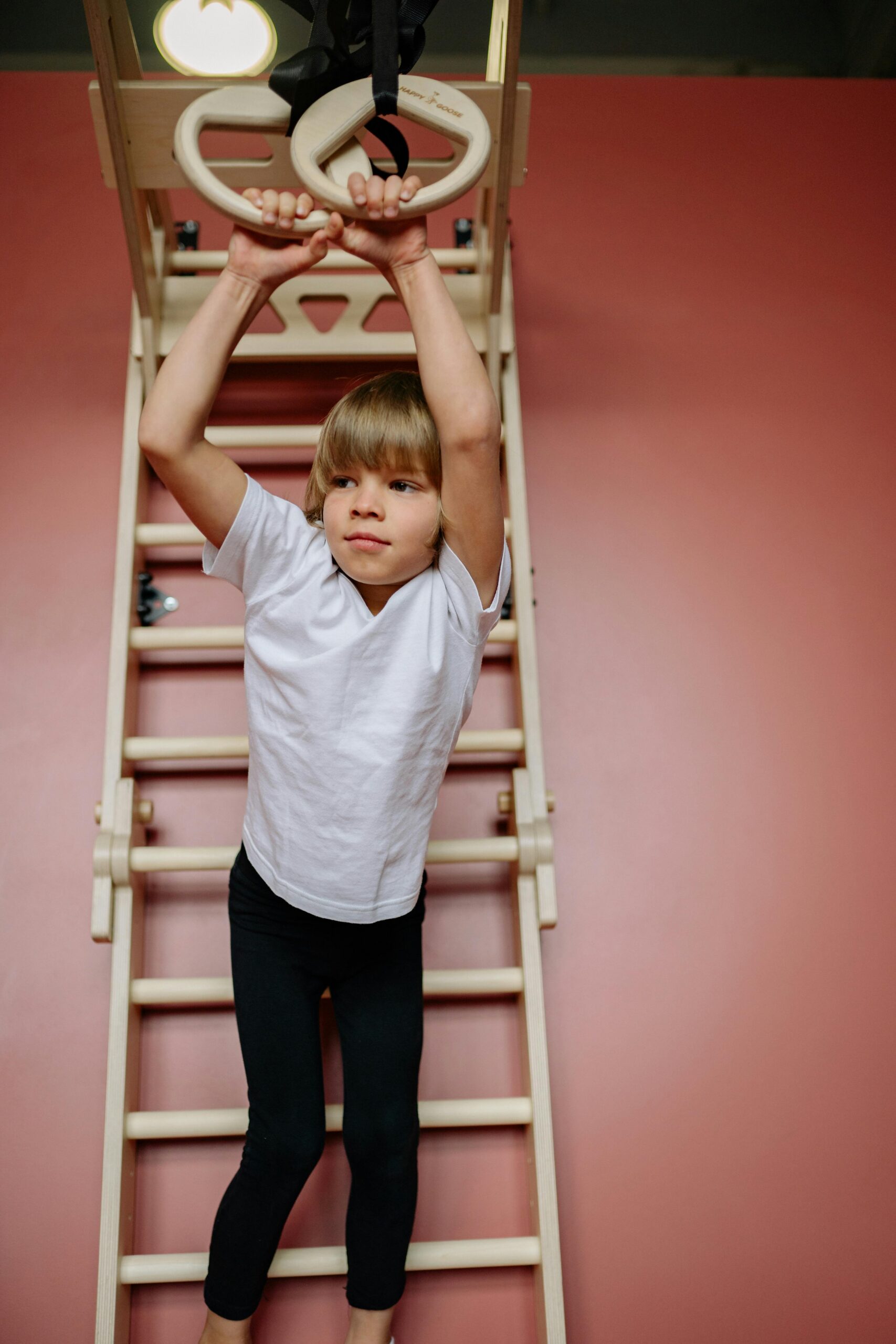 Child with blond hair hangs on gymnastic rings indoors on a wooden ladder.